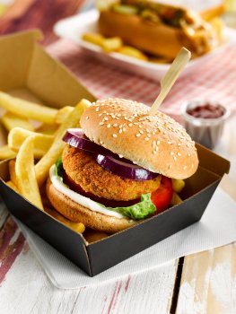 Food photograph of a breadcrumbed vegetarian burger with lettuce, tomato, mayonnaise and sliced red onion on a sesame seed bun, in a takeaway container with fries and ketchup, a hotdog and fries in the background