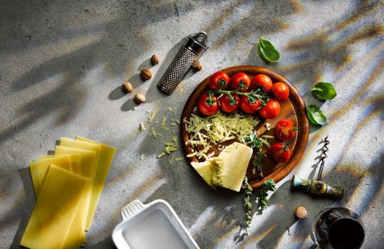Aerial food photograph of a spread of ingredients to make a beef lasagna, including pasta sheets, fresh tomatoes, red wine and grated cheese. Shot on a dark grey background with dappled light