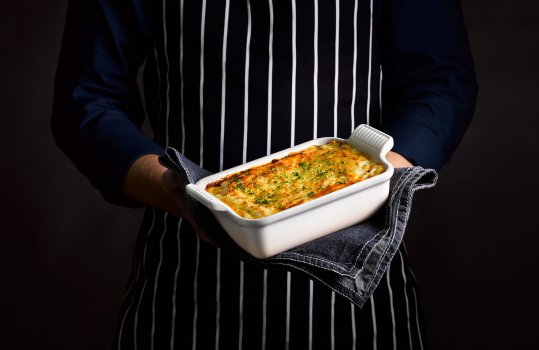 Food photograph of a man’s torso, holding a two portion dish of golden bubbling lasagna, with a golden crispy cheese top sprinkled with parsley