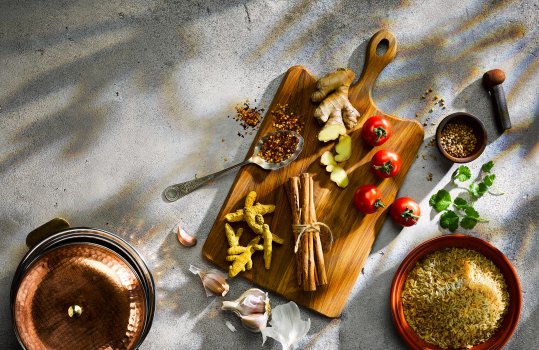 Aerial food photograph of a spread of ingredients to make a chicken makhani, including rice, ginger, garlic, turmeric and dried spices. Shot on a dark grey background with dappled light