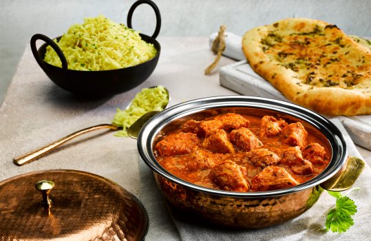 Food photograph of a two portion dish of rich chicken makhani curry, with large pieces of chopped chicken breast in a rich glossy tomatoey sauce alongside naan bread and a bowl of steamed rice
