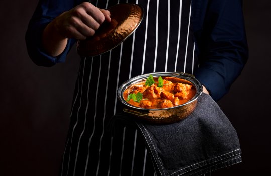 Food photograph of a man’s torso, holding a two portion dish of rich chicken makhani curry, with large pieces of chopped chicken breast in a rich glossy tomatoey sauce