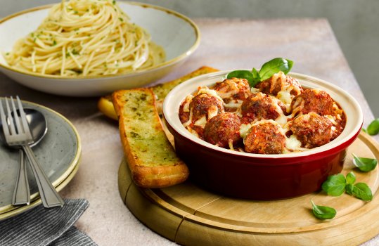 Food photograph of a two portion dish of spicy meatballs, with large spicy pork and beef meatballs in a rich glossy tomatoey sauce alongside a bowl of buttery spaghetti and garlic bread