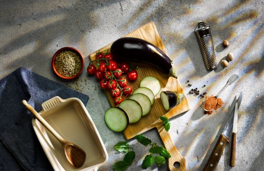 Aerial food photograph of a spread of ingredients to make a lamb moussaka, including nutmeg, aubergine and tomatoes. Shot on a dark grey background with dappled light