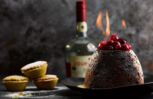 Food photograph of a large Christmas pudding topped with fresh cranberries being flambeed in brandy, served on a black platter with custard on a slate tabletop with mince pies dusted in icing sugar, and a vintage bottle of brandy in the background, with a dark wall behind