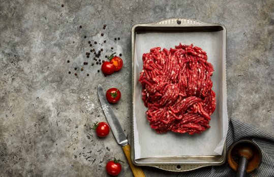 Aerial food photograph of a pile of beef mince, shot on a sheet of white parchment paper inside a vintage roasting tin, shot on a dark grey background with scattered cherry tomatoes and black peppercorns
