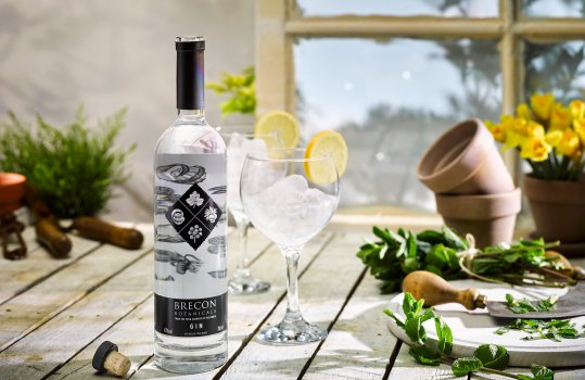 Drinks photograph of a bottle of Brecon Botanicals Gin, the bottle with the cork removed shown in a potting shed alongside gardening tools and plant cuttings, and two balloon gin glasses filled with gin, ice and lemon
