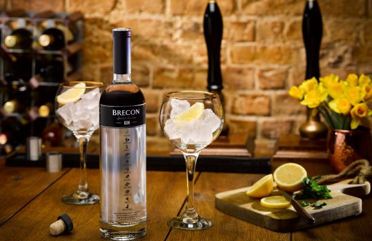 Drinks photograph of a bottle of Brecon Gin, the bottle with the cork removed shown in a bar setting with beer taps and wine racks, and two balloon gin glasses filled with, ice and lemon