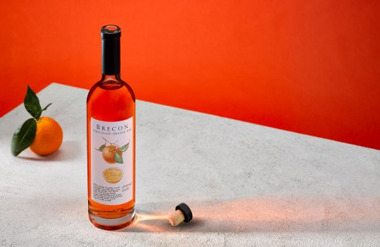 Drinks photograph of a bottle of Brecon Chocolate Orange Gin, a bottle of gin with the cork removed shown in an abstract setting on a concrete tabletop with an orange background, the same colour as the gin, with an orange with its leaves attached in the background