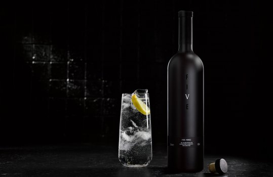 Drinks photograph of a bottle of Five vodka alongside a glass of Five vodka and tonic ice and lemon wedges. Shot in a dark atmospheric bar setting to emphasise the minimal dark branding on the bottle