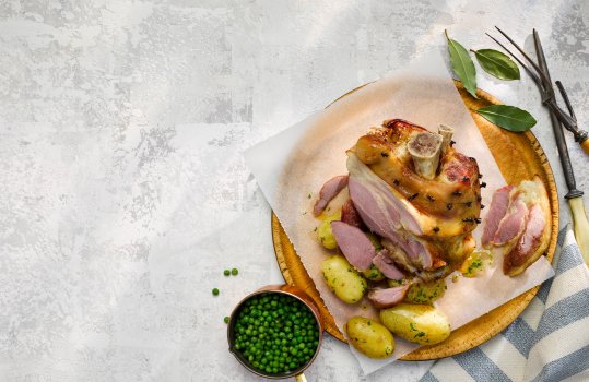 Aerial food photograph of a whole roasted gammon hock, with crispy crackling studded with cloves, shown on a wooden board alongside baby herbed potatoes and minted peas