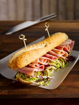 Food photograph of a ham salad baguette, a seeded baguette filled with shredded lettuce and slices of ham curled over the top. Served on baking paper on a wooden table with a chefs knife, and a white wooden wall in the background
