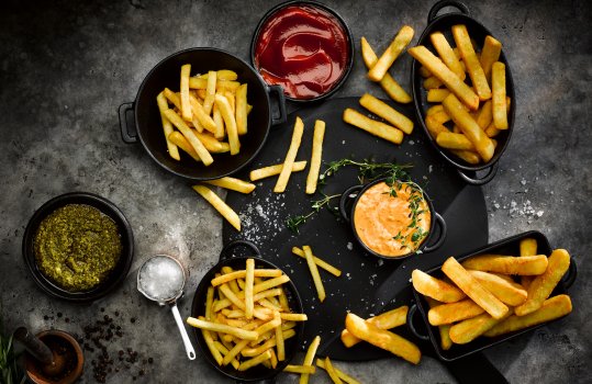 Aerial food photograph of several servings of various kinds of crispy golden chips, sprinkled with sea salt and garnished with thyme sprigs, shown on a black wooden board and black ceramic dishes, alongside black ramekins of tomato ketchup, and green pesto, in a dark atmospheric set