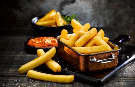 Food photograph close up of a pile of crispy golden extra thick cut chips, sprinkled with sea salt and garnished with rosemary and thyme sprigs, shown on a black wooden board in a dark atmospheric set with a black ramekin of red pesto