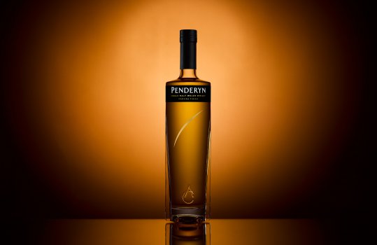 Minimalist drinks photograph of a bottle of Penderyn Madeira Finish Welsh Gold whisky, the bottle shown on a mirrored surface in front of an abstract gold background, to emphasise the rich colour of the whisky