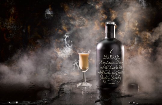 Drinks photograph of a bottle and schooner glass of Merlyn Welsh Cream Liqueur, shown in an atmospheric set on top of a black stone tabletop, in front of a crackled silver and black background with swirling smoke