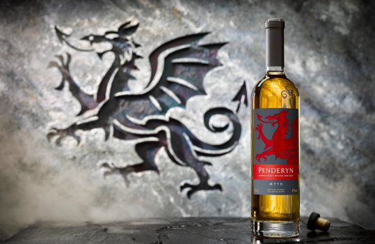 Drinks photograph of a bottle of Penderyn Myth whisky alongside a glass having been poured from the bottle, shot in a medieval looking stone set, with a carving in the stone to mirror the dragon on the label of the bottle