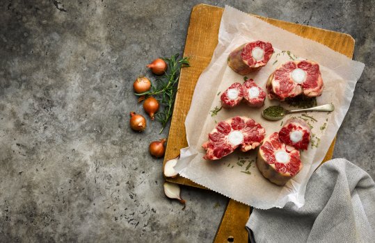 Aerial food photograph of seven pieces of raw Welsh oxtail, shown on a sheet of white parchment paper alongside fresh rosemary and shallots, and a spoon filled with dried sage