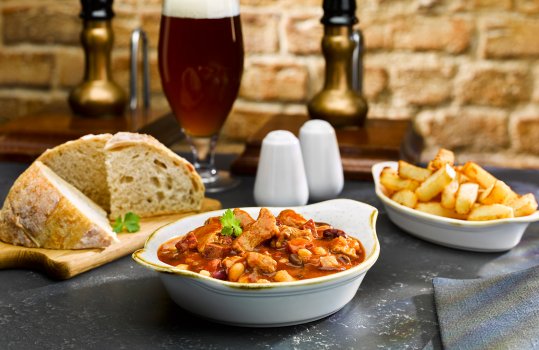 Food photograph close up of a pub style pork cassoulet, served in a white bowl alongside a dish of hand cut chips and a loaf of crusty bread. Shown in a pub setting with beer taps and a pint of dark beer in the background