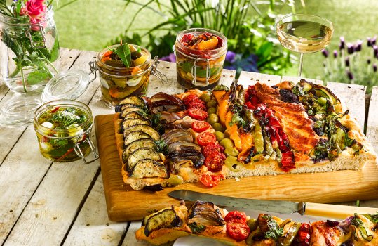 Food photograph of Spanish style foccacia, coca bread. Large tray bread topped with various Mediterranean vegetables, olives, manchego cheese and caramelised onions. Served in an outdoor setting on a wooden board on top of a white wooden table with vases of flowers, and jars of preserved vegetables in oil
