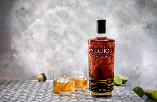 Drinks photograph of a bottle of Siddiqui Brown Rum alongside two glasses of rum and ice, with sliced limes shown in the background shot on an industrial steel background to reflect the oil rigging origins of the rum