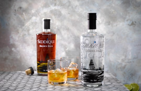 Drinks photograph of a bottle of Siddiqui White Rum alongside two glasses of rum and ice, with sliced limes and a bottle of Siddiqui Brown Rum shown in the background shot on an industrial steel background to reflect the oil rigging origins of the rum