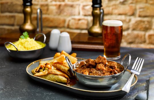 Food photograph close up of a pub style curry, a beef madras served in a silver baltic dish alongside a small ramekin of steamed rice, hand cut chips and crispy fried samosas