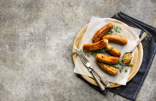 Aerial food photograph of six thick Welsh pork ‘Welsh Dragon’ sausages, cooked perfectly golden and crisp. Shown on a sheet of white parchment paper on a round wooden board alongside a spoonful of coarse wholegrain mustard