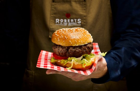 Close up food photograph of a perfect beef burger, a thick beef patty sitting on a bed of lettuce, tomato and red onion and topped with a golden sesame seed bun. Shot in the hands of a young man in a brown apron