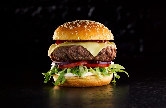 Close up food photograph of a perfect beef burger, a thick beef patty topped with melting cheese, sitting on a bed of lettuce, tomato and red onion and topped with a golden sesame seed bun. Shot on a black slate background