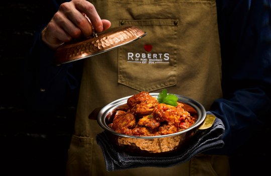 Close up food photograph of rich glossy chicken curry, with large pieces of chicken coated in deep red sauce, inside a copper dish with a lid. Shot in the hands of a young man in a brown apron