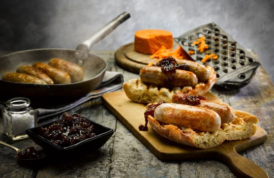 Close up food photograph of the ultimate sausage sandwich, a crisp toasted ciabatta split and filled with golden Cumberland sausages, red onion chutney and red Leicester cheese, with a frying pan of sizzling sausages, a bowl of chutney and a cheese grater in the background