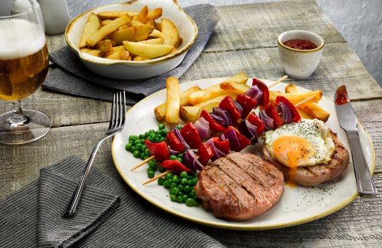Close up food photograph of a plate of homemade gammon steak, fried egg with golden liquid yolk, peas, homemade chips and vegetable skewers of red pepper and red onion. Shot on a grey background with a glass of beer