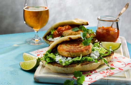 Close up food photograph of an Indian inspired chicken burger, two pan fried golden chicken burger patties with cucumber raita, lettuce, coriander, mango chutney and fresh lime inside folded naan breads, served on a white wooden board on a bright blue background