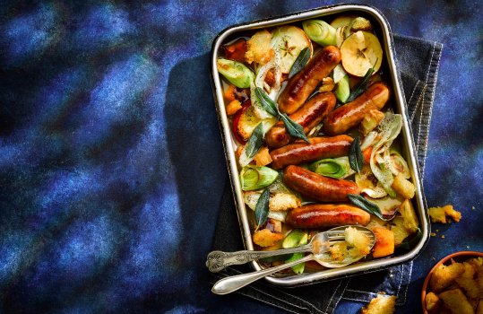 Aerial food photograph of a sausage and vegetable traybake, six golden pork and leek sausages nestled in a baking dish amongst slices of fennel, apples, leeks, butternut squash and crispy sage leaves, shot on a dark blue background