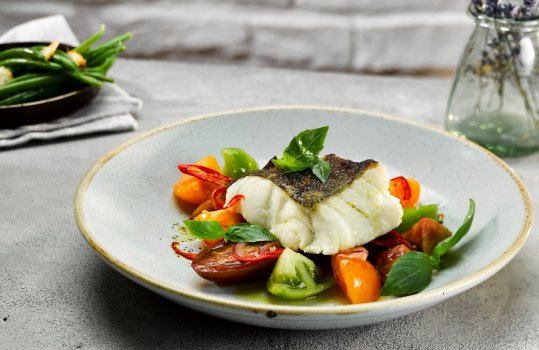 Food photograph of a juicy flakey roasted cod fillet, on top of a salad of heirloom tomatoes, chilli and basil, served in a pale blue bowl on a grey tabletop, with a pan of garlic green beans and a small glass vase of lavender in the background