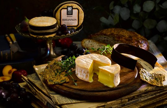 Close up food photograph of Rhiannon, an artisan cheese washed in cider and wrapped in spruce bark, one cheese is shown unwrapped and sliced on a board while there are two more in their packaging on a cake stand in the background. Shot in a dark home style set with antique books and newspapers, as well as cheeseboard accompaniments such as grapes and figs