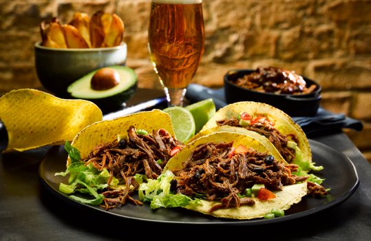Close up food photograph of a plate of homemade shredded beef tacos; lettuce, tomato salsa and shredded beef and black beans in Mexican sauce inside corn tortillas with fresh lime wedges. Shot on a dark set with a brick wall, alongside fresh avocado, extra corn tortillas and a glass of beer
