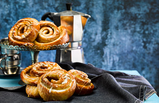 Food photograph close up of a pile of cinnamon Danish pastries drizzles with icing. A pile of pastries in the foreground with more shown on a cake stand in the background, next to a stack of coffee cups and a coffee pot against a dark blue background