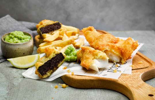 Food photograph of fish and chips, a crispy battered flakey cod fillet alongside homemade french fries, and a slice of crispy battered black pudding, served on a wooden board alongside mushy peas on a grey tabletop with a grey wall in the background