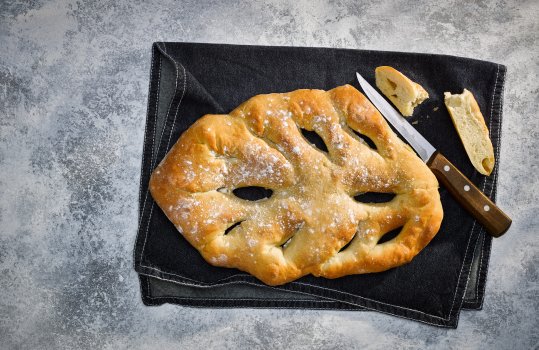 Aerial food photograph of a homemade fougasse bread, traditionally shaped French bread in the shape of a feather or leaf, shown with slices removed on a navy blue tea towel