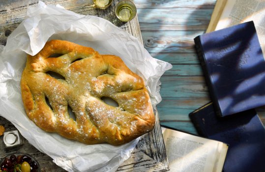 Aerial food photograph of a homemade fougasse bread, traditionally shaped French bread in the shape of a feather or leaf, shown alongside marinated olives and glasses of Pastisse on white baking paper