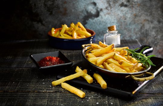 Food photograph close up of a pile of crispy golden skin on medium cut chips, sprinkled with sea salt and garnished with rosemary sprigs, shown on a black wooden board in a dark atmospheric set with a black ramekin of tomato chutney