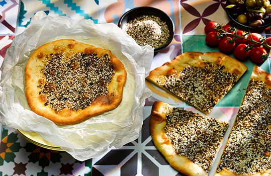 Aerial food photograph of a homemade man’ouche, a traditional Levantine flatbread topped with za’atar, shot on a vibrant tiled background alongside olives, tomatoes and a small ramekin of za’atar