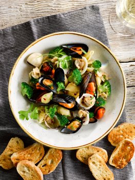 Aerial food photograph of clam and mussel pasta with broccoli florets and parsley, served with golden toasted baguette and a glass of white wine
