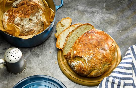 Aerial food photograph of two loaves of homemade no knead bread, one shown sliced open on a wooden cutting board and the other inside a cast iron dutch oven lined with baking parchment