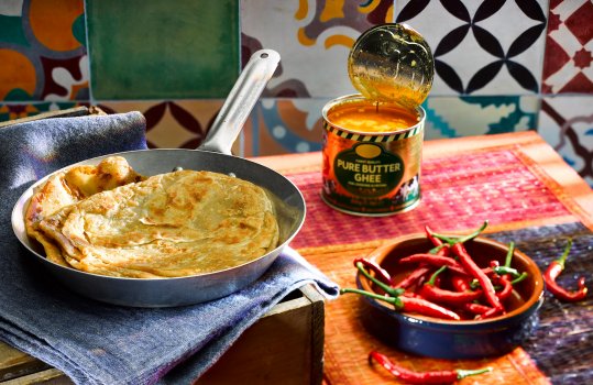 Food photograph of a pan of homemade parathas, a vintage frying pan with a paratha inside it on a blue tea towel, shot on a vibrant tiled background alongside a ramekin of chillies and an open tin of ghee for brushing onto the parathas