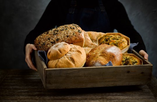 Food photograph of a woman in a black jumper and denim apron holding a wooden crate filled with loaves of homemade bread