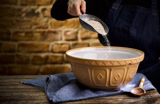 Food photograph of a woman in a black jumper and denim apron sprinkling flour from a large metal scoop into a vintage beige mixing bowl