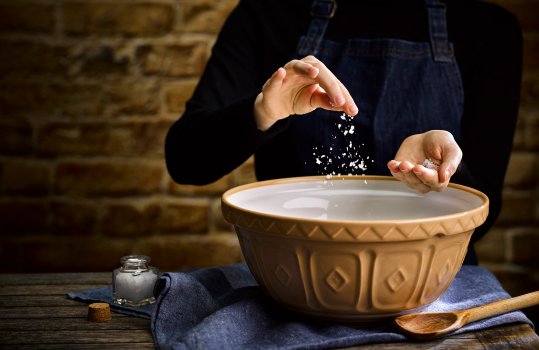 Food photograph of a woman in a black jumper and denim apron sprinkling flakes of sea salt into a vintage beige mixing bowl